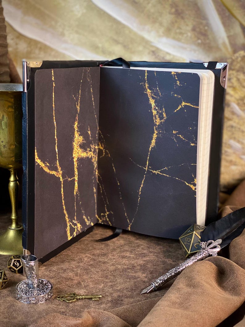 Puede incluir: Cuaderno negro abierto con vetas doradas en las p&aacute;ginas interiores. El cuaderno tiene una cinta negra como marcador y protectores de esquina met&aacute;licos. Un bol&iacute;grafo plateado, una llave y dados est&aacute;n sobre la superficie marr&oacute;n.