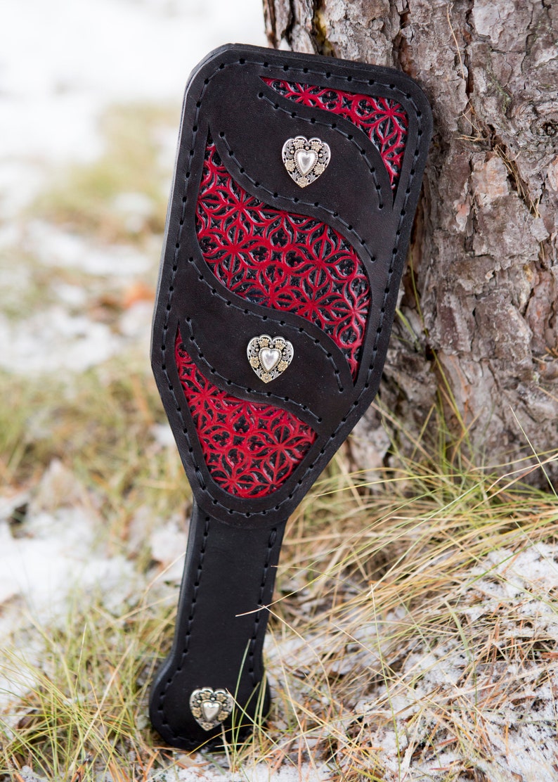 May include: A black leather paddle with red and black floral detailing and three silver heart-shaped accents.