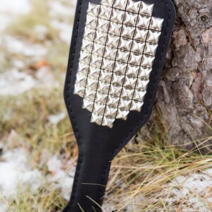 May include: A black leather paddle with silver pyramid-shaped studs. The paddle is lying on a bed of brown grass and snow.