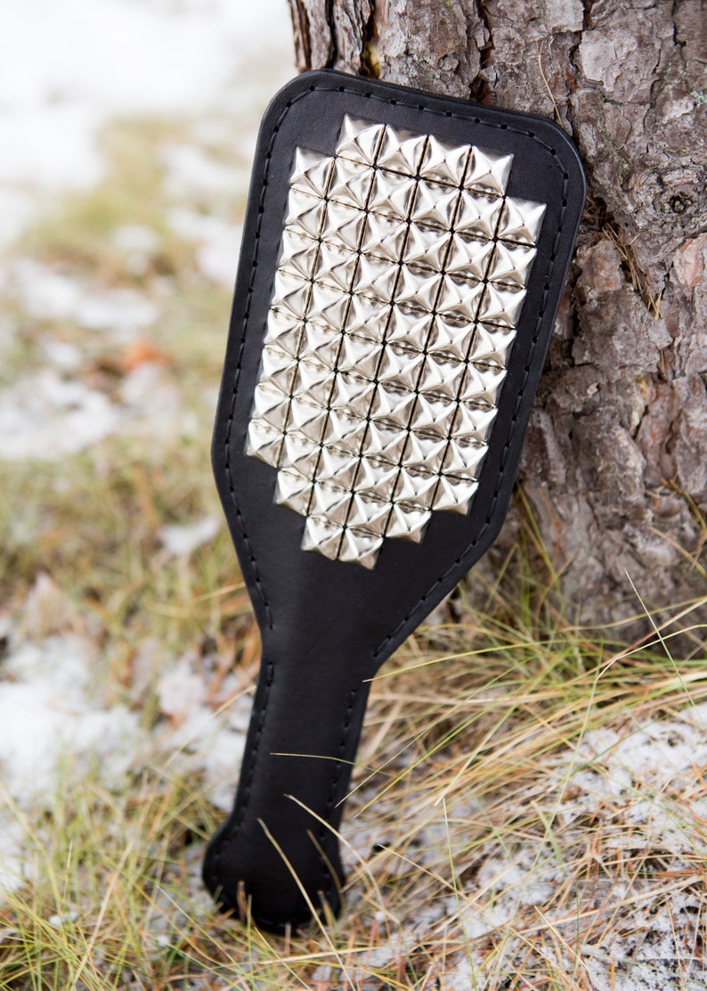 May include: A black leather paddle with silver pyramid-shaped studs. The paddle is lying on a bed of brown grass and snow.