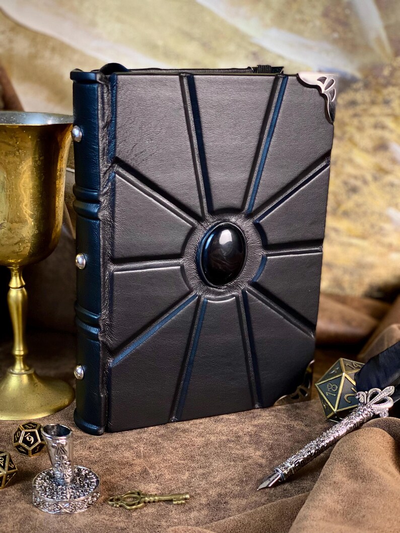 Puede incluir: Diario de cuero negro con un dise&ntilde;o geom&eacute;trico y una piedra negra central. El diario tiene detalles plateados en las esquinas y un lomo azul oscuro con remaches plateados. Un c&aacute;liz dorado y dados en el fondo.