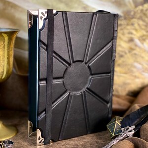 Puede incluir: Diario de cuero negro con dise&ntilde;o de sol en relieve en la portada. Cuenta con un cierre de banda el&aacute;stica negra y protectores de esquina met&aacute;licos. Tambi&eacute;n se ven un c&aacute;liz dorado, una pluma y un tintero decorativo.