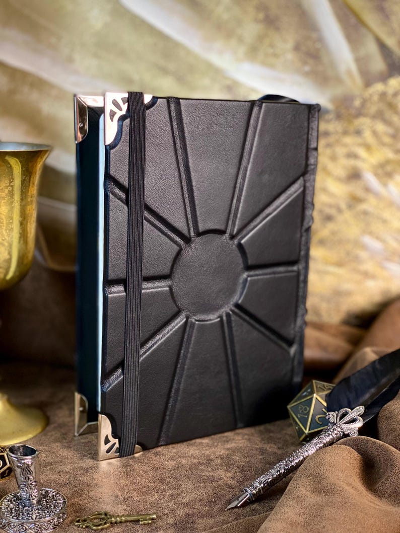 Puede incluir: Diario de cuero negro con dise&ntilde;o de sol en relieve en la portada. Cuenta con un cierre de banda el&aacute;stica negra y protectores de esquina met&aacute;licos. Tambi&eacute;n se ven un c&aacute;liz dorado, una pluma y un tintero decorativo.