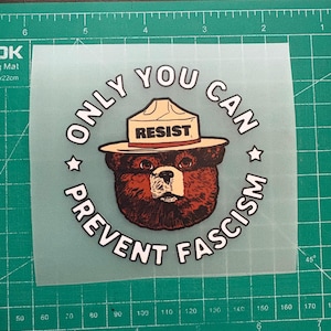 May include: A circular sticker featuring a brown bear wearing a ranger hat with the word "RESIST" on it. The text "ONLY YOU CAN PREVENT FASCISM" surrounds the bear image in a circular pattern.