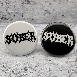 May include: Two circular pin-back buttons featuring the word SOBER in a stylized, jagged black-letter font. One button is white with black text, and the other button is black with white text. Both buttons have a smooth, glossy finish.