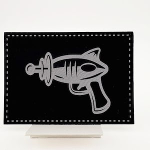 May include: A black and white illustration of a ray gun on a black background with a white stitched border.