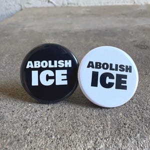 May include: Two round buttons, one black and one white, each with the words "ABOLISH ICE" in bold, white lettering. The buttons are displayed on a textured, gray surface.