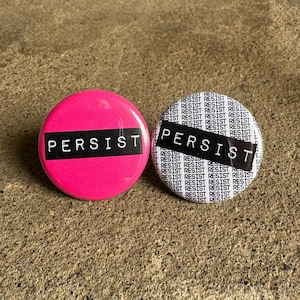 May include: Two round button pins with the word "PERSIST" printed in black on a white background and a pink background. The white button pin has the word "RESIST" repeated in black text on a white background.