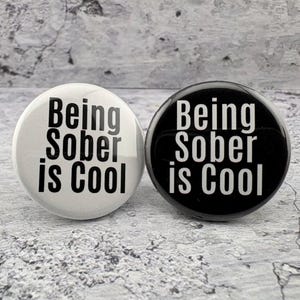 May include: Two circular pin-back buttons featuring the text Being Sober is Cool. One button is white with black text, and the other button is black with white text. Both buttons have a glossy finish and a simple, bold sans-serif font design.