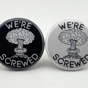 May include: Two round buttons with the text "We're Screwed" and a black and white illustration of an exploding mushroom cloud. One button is black with white text and illustration, the other is white with black text and illustration.