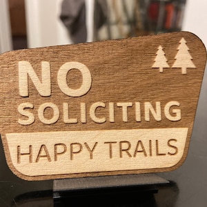 May include: Wooden sign with the words "NO SOLICITING" and "HAPPY TRAILS" engraved in a light color. Two small tree silhouettes are in the upper right corner. The sign is mounted on a dark metal stand.