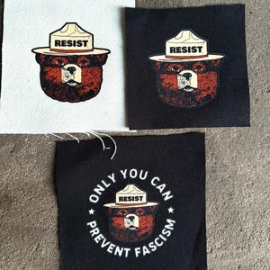 May include: Three square patches featuring a bear wearing a ranger hat with the word "RESIST" on it. One patch is on white fabric, one on black, and the third patch on black fabric has the text "ONLY YOU CAN PREVENT FASCISM".
