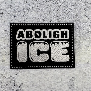 May include: A black rectangular patch with the words "ABOLISH ICE" in bold, silver lettering. The letters are outlined in a lighter silver color, and the patch is bordered with white stitching. The background is a textured, light gray surface.