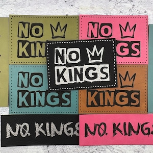 May include: A collection of rectangular patches in various colors, including black, pink, teal, and brown. Each patch features the words "NO KINGS" in bold, stylized lettering, with a crown graphic above the words. The patches are likely for clothing or accessories.