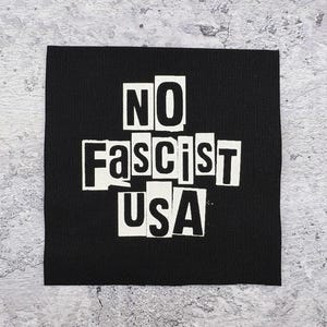 May include: Black square patch with the words "NO Fascist USA" in white, block-style letters. The text is arranged in a cross shape. The patch is made of a textured fabric.