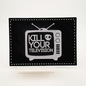 May include: Black leather patch with a white stitched border. The patch features a white silhouette of a vintage television with the text "KILL YOUR TELEVISION" and a skull graphic.