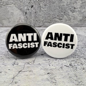May include: Two round buttons with the words "ANTI FASCIST" in bold, white letters. One button is black, the other is white. The buttons are on a textured, gray surface.