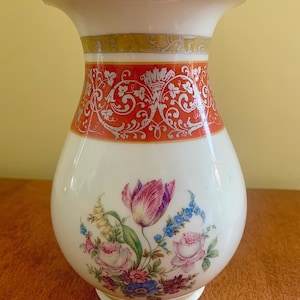 May include: A white ceramic vase featuring a wide, flared rim and a rounded body. The neck is decorated with a horizontal band of orange and silver filigree patterns. The lower body displays a colorful floral bouquet of tulips, roses, and blue flowers.