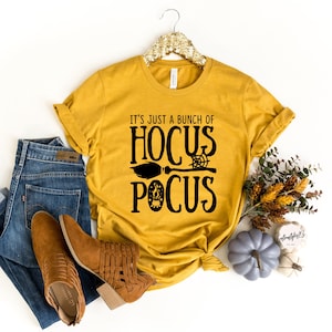 Pode incluir: Uma camiseta amarela mostarda com texto preto que diz "It's just a bunch of Hocus Pocus" com uma vassoura e outros gr&aacute;ficos tem&aacute;ticos de Halloween.