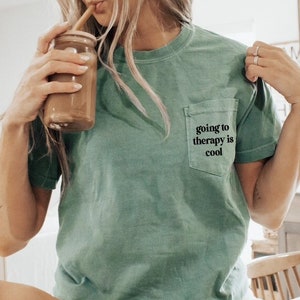 May include: A woman wearing a green t-shirt with a pocket that says "going to therapy is cool" and white socks with black stripes. She is sitting on a white surface and holding a glass with a straw.