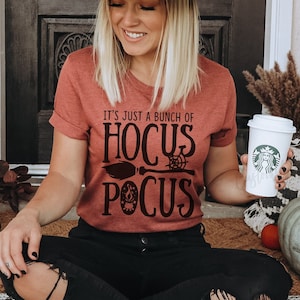 Pode incluir: Uma mulher usando um chap&eacute;u preto com detalhes prateados e uma camiseta cor ferrugem com o texto "It's just a bunch of Hocus Pocus" e um gr&aacute;fico de vassoura preta e teia de aranha. Ela est&aacute; segurando um copo branco para viagem com um logotipo preto.