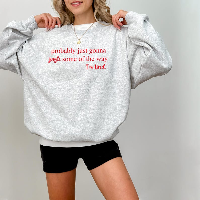 May include: A light gray sweatshirt with the text "probably just gonna jingle some of the way I'm tired" in red. The sweatshirt is paired with black shorts. The model is wearing a necklace.