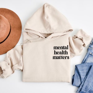 May include: A beige hoodie with a black graphic that says "mental health matters". The hoodie is laid out on a white surface with a brown hat and blue jeans.