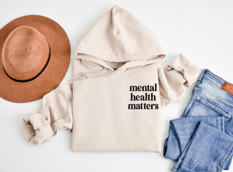 May include: A beige hoodie with a black graphic that says "mental health matters". The hoodie is laid out on a white surface with a brown hat and blue jeans.