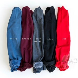 May include: Five folded sweatshirts in different colours: indigo, maroon, dark heather, black, and red. Each sweatshirt has the colour name printed on the cuff.