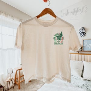 Pode incluir: Uma t-shirt de manga curta cor de creme com um gr&aacute;fico na parte superior esquerda do peito. O gr&aacute;fico apresenta um emblema de &aacute;guia verde estilizado com detalhes em vermelho e verde, posicionado acima da palavra MEXICO em letras verdes.