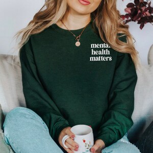 May include: A woman wearing a dark green sweatshirt with the text "mental health matters" in white lettering. She is sitting on a sofa with a white and grey cushion behind her.
