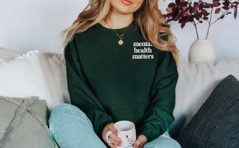 May include: A woman wearing a dark green sweatshirt with the text "mental health matters" in white lettering. She is sitting on a sofa with a white and grey cushion behind her.