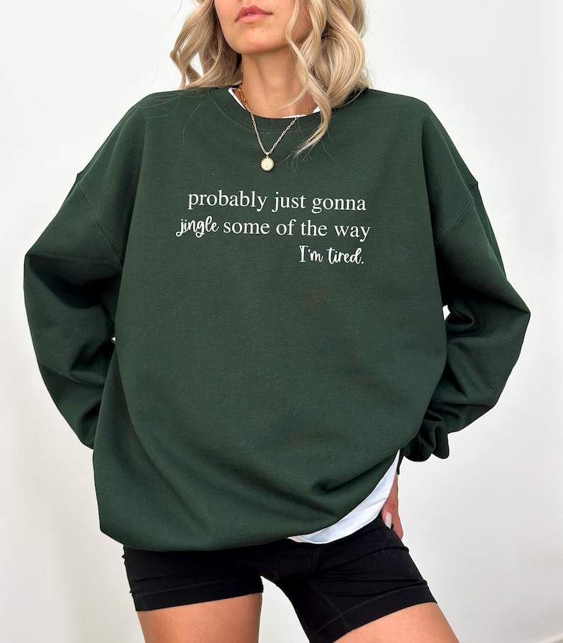 May include: A dark green sweatshirt with the text "probably just gonna jingle some of the way I'm tired." The sweatshirt has long sleeves and a white undershirt peeking out at the neckline and hem. The person is wearing black shorts.