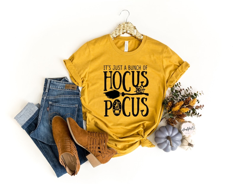 Pode incluir: Uma camiseta amarela mostarda com texto preto que diz "It's just a bunch of Hocus Pocus" com uma vassoura e outros gr&aacute;ficos tem&aacute;ticos de Halloween.