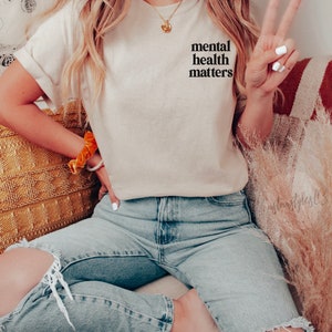 May include: A woman wearing a light beige t-shirt with the text "mental health matters" printed on the front. She is wearing blue jeans with a distressed knee and is sitting on a sofa.