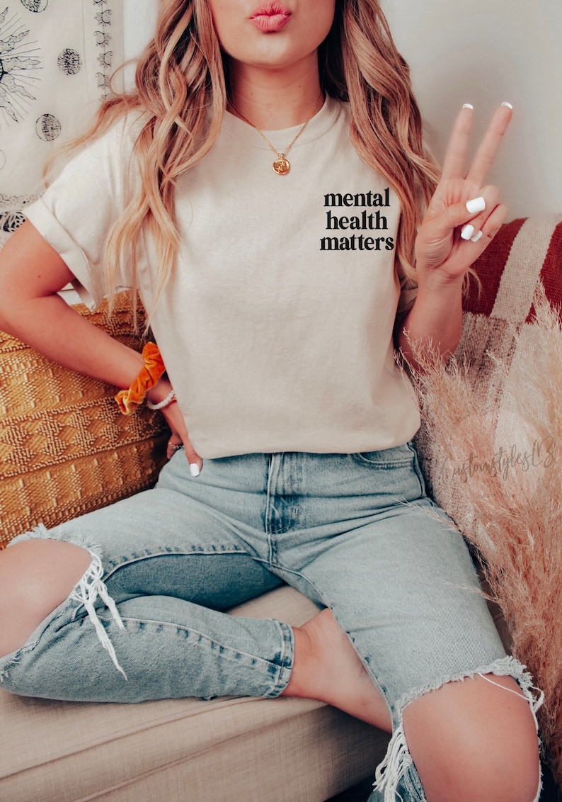 May include: A woman wearing a light beige t-shirt with the text "mental health matters" printed on the front. She is wearing blue jeans with a distressed knee and is sitting on a sofa.