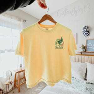 Pode incluir: Uma t-shirt de manga curta em amarelo p&aacute;lido com um gr&aacute;fico no lado esquerdo do peito. O design inclui um emblema de &aacute;guia verde dentro de uma borda circular com detalhes vermelhos e a palavra MEXICO em letras mai&uacute;sculas verdes.