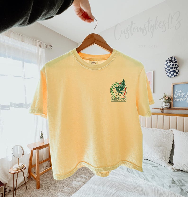 Pode incluir: Uma t-shirt de manga curta em amarelo p&aacute;lido com um gr&aacute;fico no lado esquerdo do peito. O design inclui um emblema de &aacute;guia verde dentro de uma borda circular com detalhes vermelhos e a palavra MEXICO em letras mai&uacute;sculas verdes.