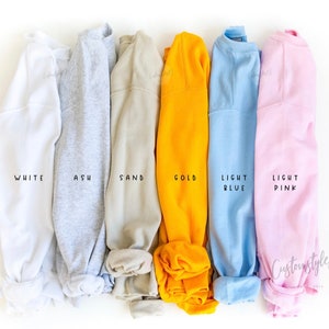May include: Six folded sweatshirts in different colours: white, ash, sand, gold, light blue, and light pink. The colours are labelled below each sweatshirt.