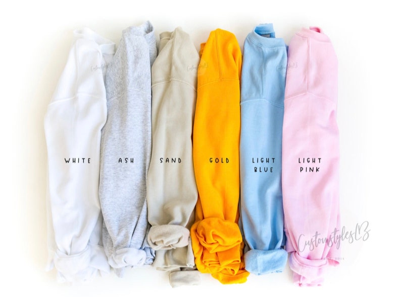 May include: Six folded sweatshirts in different colours: white, ash, sand, gold, light blue, and light pink. The colours are labelled below each sweatshirt.