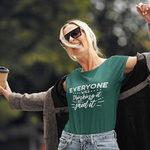 Puede incluir: Una mujer con una camiseta verde con el texto "Everyone was thinking it... I just said it." Ella sostiene una taza de papel marr&oacute;n y sonr&iacute;e.