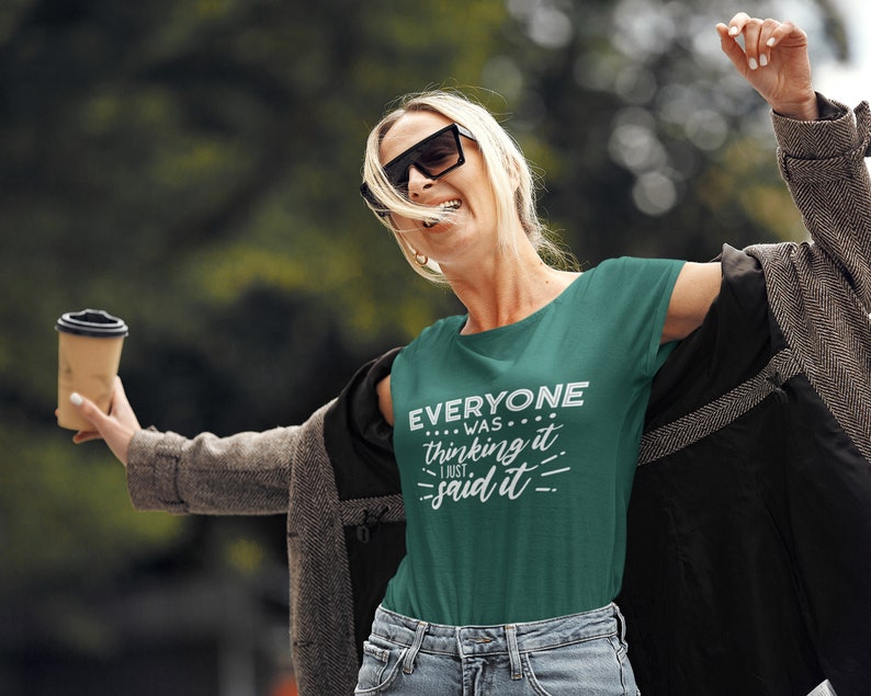 Puede incluir: Una mujer con una camiseta verde con el texto "Everyone was thinking it... I just said it." Ella sostiene una taza de papel marr&oacute;n y sonr&iacute;e.