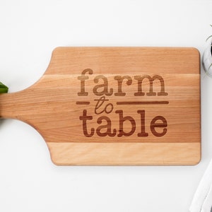 May include: A wooden cutting board with a handle, featuring the text "farm to table" engraved on the surface.