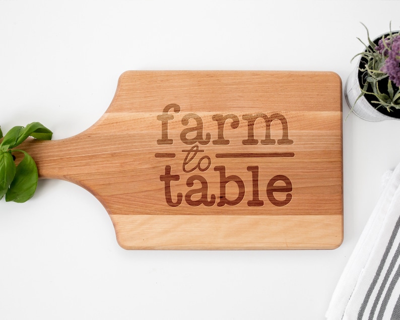 May include: A wooden cutting board with a handle, featuring the text "farm to table" engraved on the surface.