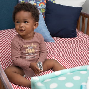 Pu&ograve; includere: Un bambino piccolo che indossa una maglietta a maniche lunghe color marrone con il testo "Thou shall not tame me - Toddler 24/7" in giallo. Il bambino &egrave; seduto in una culla con lenzuola a righe rosse e bianche.