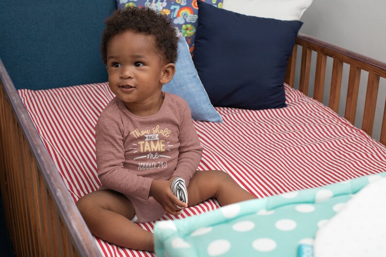 Pu&ograve; includere: Un bambino piccolo che indossa una maglietta a maniche lunghe color marrone con il testo "Thou shall not tame me - Toddler 24/7" in giallo. Il bambino &egrave; seduto in una culla con lenzuola a righe rosse e bianche.