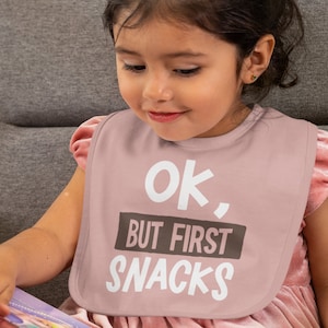 Pu&ograve; includere: Un bavaglino rosa per neonati con il testo "Ok, But First Snacks" in lettere bianche e marroni.