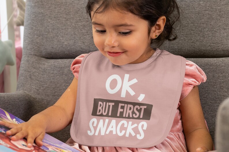 Pu&ograve; includere: Un bavaglino rosa per neonati con il testo "Ok, But First Snacks" in lettere bianche e marroni.