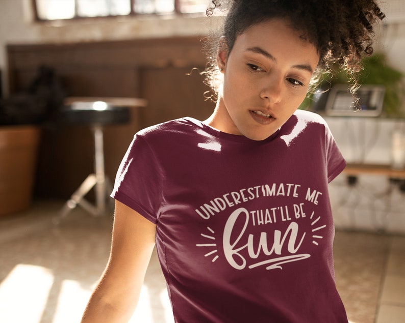 Puede incluir: Una camiseta color granate con un gr&aacute;fico blanco que dice "Underestimate me that'll be fun".