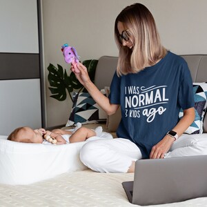 Pode incluir: Uma mulher vestindo uma camiseta azul marinho com o texto "Eu era normal h&aacute; 3 filhos" est&aacute; sentada em uma cama com um laptop. Um beb&ecirc; est&aacute; deitado em um ber&ccedil;o branco e brincando com um brinquedo roxo.
